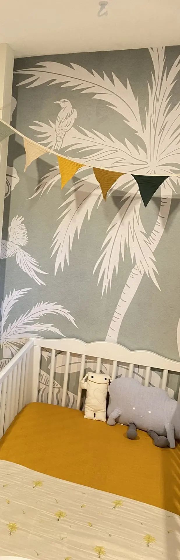 Fresque jungle sauge monochrome et fanions au-dessus d’un lit de bébé.